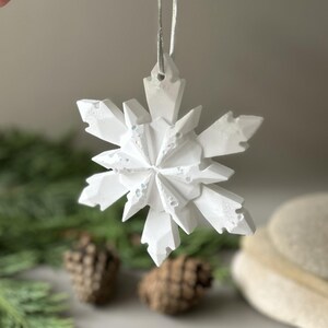 Pode incluir: Um ornamento de floco de neve branco com m&uacute;ltiplas facetas, acabamento mate texturizado e pontas geom&eacute;tricas afiadas. O ornamento est&aacute; suspenso por uma fita fina de cor prateada presa no topo, criando um design n&iacute;tido, angular e sim&eacute;trico.