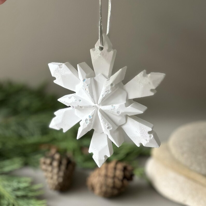 Pode incluir: Um ornamento de floco de neve branco com m&uacute;ltiplas facetas, acabamento mate texturizado e pontas geom&eacute;tricas afiadas. O ornamento est&aacute; suspenso por uma fita fina de cor prateada presa no topo, criando um design n&iacute;tido, angular e sim&eacute;trico.