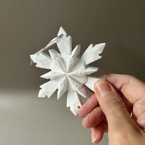 Pode incluir: Um ornamento de floco de neve branco de v&aacute;rias pontas com um design geom&eacute;trico facetado. A superf&iacute;cie apresenta manchas iridescentes subtis incrustadas que captam a luz. Uma fita fina de cor prateada est&aacute; presa no topo para pendurar.