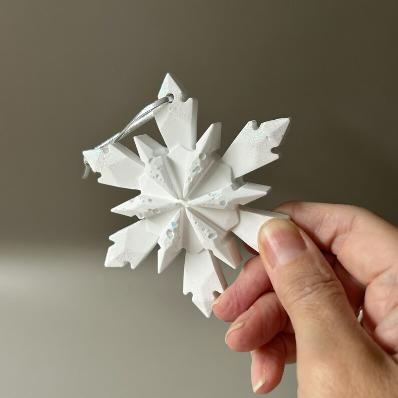 Pode incluir: Um ornamento de floco de neve branco de v&aacute;rias pontas com um design geom&eacute;trico facetado. A superf&iacute;cie apresenta manchas iridescentes subtis incrustadas que captam a luz. Uma fita fina de cor prateada est&aacute; presa no topo para pendurar.