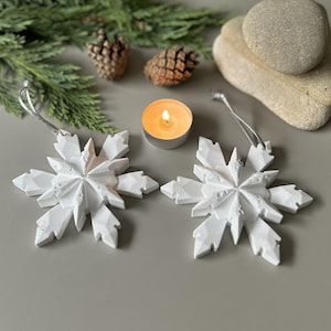 Pode incluir: Dois ornamentos de floco de neve brancos com m&uacute;ltiplas facetas e cord&otilde;es prateados para pendurar. Cada floco de neve apresenta um design geom&eacute;trico em camadas com subtis reflexos de glitter iridescente na superf&iacute;cie, criando um aspeto texturado.