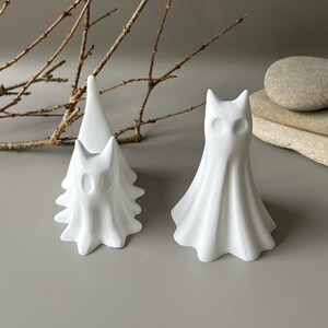 May include: Two white matte ceramic ghost figurines with smooth, flowing drapery textures and hollow oval eye sockets. One figurine is taller and stands upright, while the other is slightly smaller and positioned behind it, creating a layered effect.