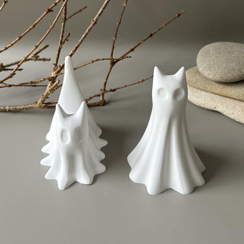 May include: Two white matte ceramic ghost figurines with smooth, flowing drapery textures and hollow oval eye sockets. One figurine is taller and stands upright, while the other is slightly smaller and positioned behind it, creating a layered effect.