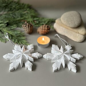 Pode incluir: Dois ornamentos geom&eacute;tricos de floco de neve brancos com subtis manchas iridescentes, cada um com um cord&atilde;o prateado para pendurar. Os ornamentos s&atilde;o exibidos ao lado de uma pequena vela acesa, duas pinhas castanhas, agulhas de pinheiro e pedras lisas.