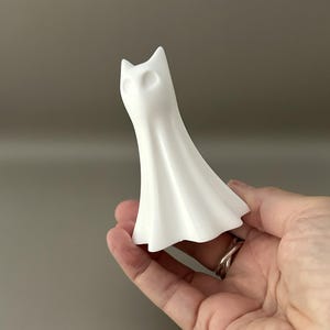 May include: A small, matte white figurine shaped like a stylised ghost with pointed ears and hollow, oval-shaped eye sockets. The object has a smooth, flowing, draped form that flares out at the base, resembling a sheet-covered figure or a cat-like spirit.