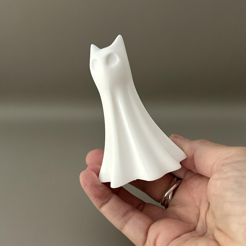 May include: A small, matte white figurine shaped like a stylised ghost with pointed ears and hollow, oval-shaped eye sockets. The object has a smooth, flowing, draped form that flares out at the base, resembling a sheet-covered figure or a cat-like spirit.