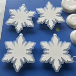 Pode incluir: Quatro moldes de silicone brancos em forma de floco de neve dispostos sobre uma superf&iacute;cie azul. Cada molde apresenta um design detalhado de estrela de seis pontas com ramos intrincados e um pequeno la&ccedil;o circular no topo para pendurar.