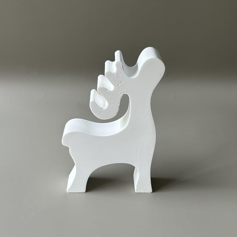 May include: A solid white, stylized reindeer figurine standing in profile. The object features a smooth, matte finish with a simplified, geometric silhouette, including a curved neck, a short tail, and distinct, rounded antler shapes.