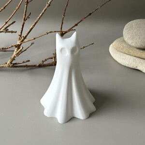 May include: A white, smooth, matte-finished figurine shaped like a ghost with pointed ears and two circular, hollowed-out eye sockets. The object has a flared, draped base that resembles fabric folds, standing upright as a singular decorative piece.