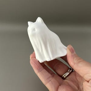 Może przedstawiać: Mała, matowobiała figurka w kształcie kota przebranego za ducha. Obiekt posiada spiczaste uszy, okrągłe wgłębienia na oczy oraz pionowe fałdy wzdłuż korpusu, przypominające prześcieradło przykrywające stojącą postać kota.
