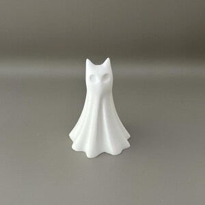 May include: A small, white, matte-finished figurine shaped like a stylised ghost with pointed ears and two hollow, circular eye sockets. The figure has a smooth, flowing, draped appearance that flares out at the base, creating a bell-like silhouette.
