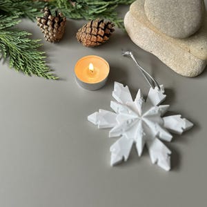 Pode incluir: Um ornamento de floco de neve branco de v&aacute;rias pontas com um cord&atilde;o prateado para pendurar. O ornamento apresenta um design geom&eacute;trico facetado com bordas afiadas e angulares. &Eacute; exibido ao lado de duas pinhas castanhas, uma vela de ch&aacute; acesa e pedras.