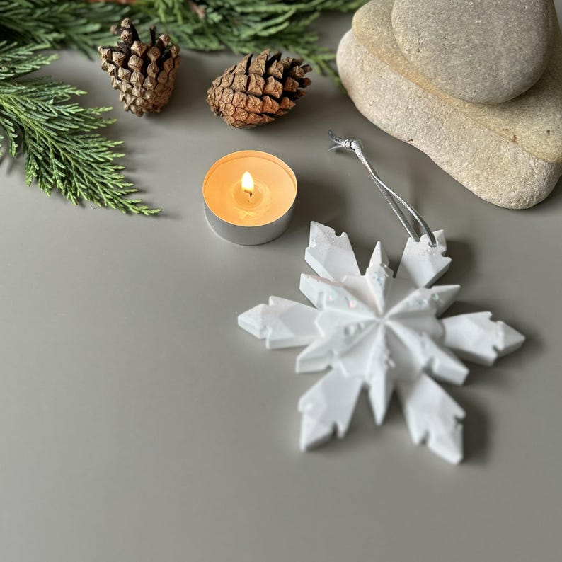 Pode incluir: Um ornamento de floco de neve branco de v&aacute;rias pontas com um cord&atilde;o prateado para pendurar. O ornamento apresenta um design geom&eacute;trico facetado com bordas afiadas e angulares. &Eacute; exibido ao lado de duas pinhas castanhas, uma vela de ch&aacute; acesa e pedras.