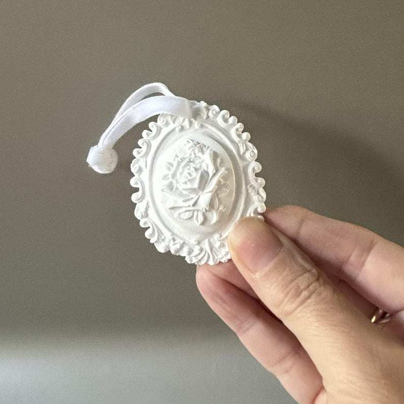 K&ouml;nnte beinhalten: Ein wei&szlig;es, ovales Ornament mit einem erhabenen Relief einer Rose in der Mitte, umgeben von einem verzierten, gewellten Rand. Das St&uuml;ck enth&auml;lt eine kleine wei&szlig;e Stoffschlaufe an der Oberseite zum Aufh&auml;ngen, die einen dekorativen Akzent setzt.