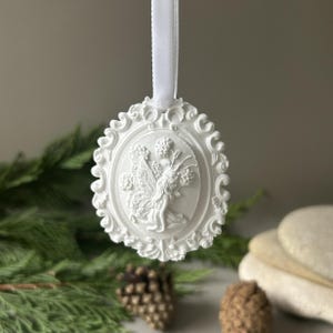 K&ouml;nnte beinhalten: Ein wei&szlig;es, ovales H&auml;ngeornament mit einem Relief einer gefl&uuml;gelten Fee, die von Blumen umgeben ist. Das Ornament hat einen verzierten, geschwungenen Rand und ist an einer passenden wei&szlig;en Satinschleife oben in der Mitte aufgeh&auml;ngt.