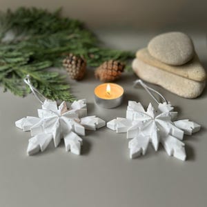 Pode incluir: Dois ornamentos geom&eacute;tricos de floco de neve brancos com cord&otilde;es prateados. Cada floco de neve apresenta um design angular facetado com acabamento fosco. Os ornamentos est&atilde;o posicionados numa superf&iacute;cie plana, acompanhados por uma vela acesa.