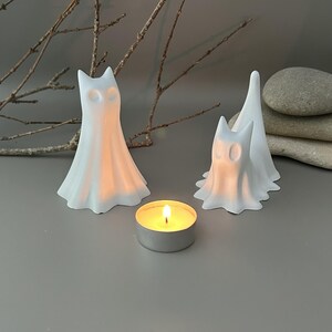 May include: Two white, ghost-shaped ceramic figurines with hollow eye sockets, positioned next to a small, round metal tea light candle holder containing a lit candle with a warm yellow flame, creating a soft glow on the smooth surface.