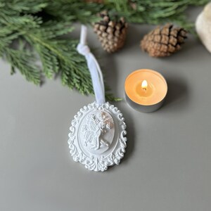 K&ouml;nnte beinhalten: Ein wei&szlig;es, ovales H&auml;ngeornament mit einem Relief einer Feenfigur, umgeben von einem verzierten, geschwungenen Rand. Das Ornament ist an einem wei&szlig;en Band befestigt und weist detaillierte Ausarbeitungen am zentralen Feenmotiv auf.