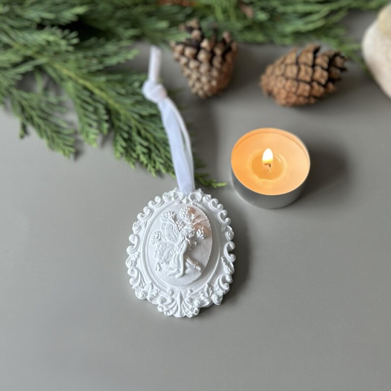 K&ouml;nnte beinhalten: Ein wei&szlig;es, ovales H&auml;ngeornament mit einem Relief einer Feenfigur, umgeben von einem verzierten, geschwungenen Rand. Das Ornament ist an einem wei&szlig;en Band befestigt und weist detaillierte Ausarbeitungen am zentralen Feenmotiv auf.