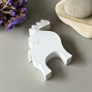 May include: A solid white, matte-finished figurine shaped like a stylized moose. The object features a smooth, flat surface with a distinct silhouette of the animal, including antlers and four legs, standing in a fixed, upright position.