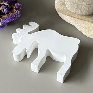 May include: A solid white, matte-finished moose figurine crafted in a flat, silhouette-style design. The object features the distinct shape of a moose with prominent antlers, a long snout, and four legs, standing as a singular, decorative piece.