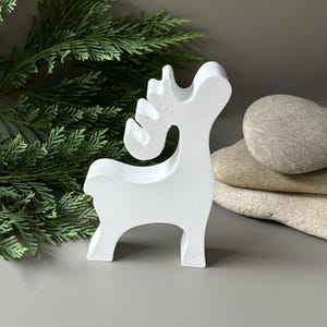 May include: A white, stylized reindeer figurine with a smooth, matte finish. The object features a simplified silhouette with a curved neck, a rounded head, and prominent antlers. The reindeer stands on four legs and is oriented to the right.
