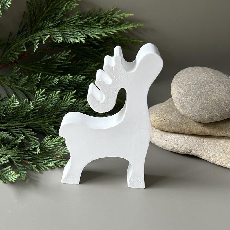 May include: A white, stylized reindeer figurine with a smooth, matte finish. The object features a simplified silhouette with a curved neck, a rounded head, and prominent antlers. The reindeer stands on four legs and is oriented to the right.