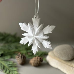 Pode incluir: Um ornamento de floco de neve branco com m&uacute;ltiplas facetas e uma estrutura geom&eacute;trica cristalina. O ornamento apresenta pontas angulares afiadas e uma superf&iacute;cie texturizada com pequenas reentr&acirc;ncias, suspenso por um fio fino de cor clara.