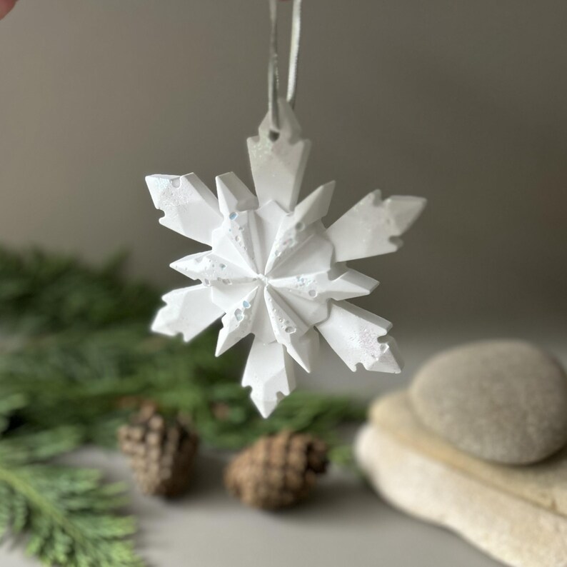 Pode incluir: Um ornamento de floco de neve branco com m&uacute;ltiplas facetas e uma estrutura geom&eacute;trica cristalina. O ornamento apresenta pontas angulares afiadas e uma superf&iacute;cie texturizada com pequenas reentr&acirc;ncias, suspenso por um fio fino de cor clara.