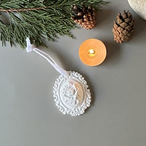 K&ouml;nnte beinhalten: Ein wei&szlig;es, ovales H&auml;ngeornament mit einem detaillierten Relief einer Rose in der Mitte, umgeben von einem verzierten, schn&ouml;rkelartigen Rand. Das Ornament ist an einer d&uuml;nnen wei&szlig;en Bandschlaufe zum Aufh&auml;ngen befestigt und hat eine matte Oberfl&auml;che.