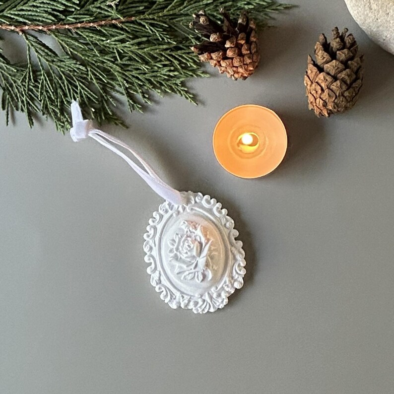 K&ouml;nnte beinhalten: Ein wei&szlig;es, ovales H&auml;ngeornament mit einem detaillierten Relief einer Rose in der Mitte, umgeben von einem verzierten, schn&ouml;rkelartigen Rand. Das Ornament ist an einer d&uuml;nnen wei&szlig;en Bandschlaufe zum Aufh&auml;ngen befestigt und hat eine matte Oberfl&auml;che.