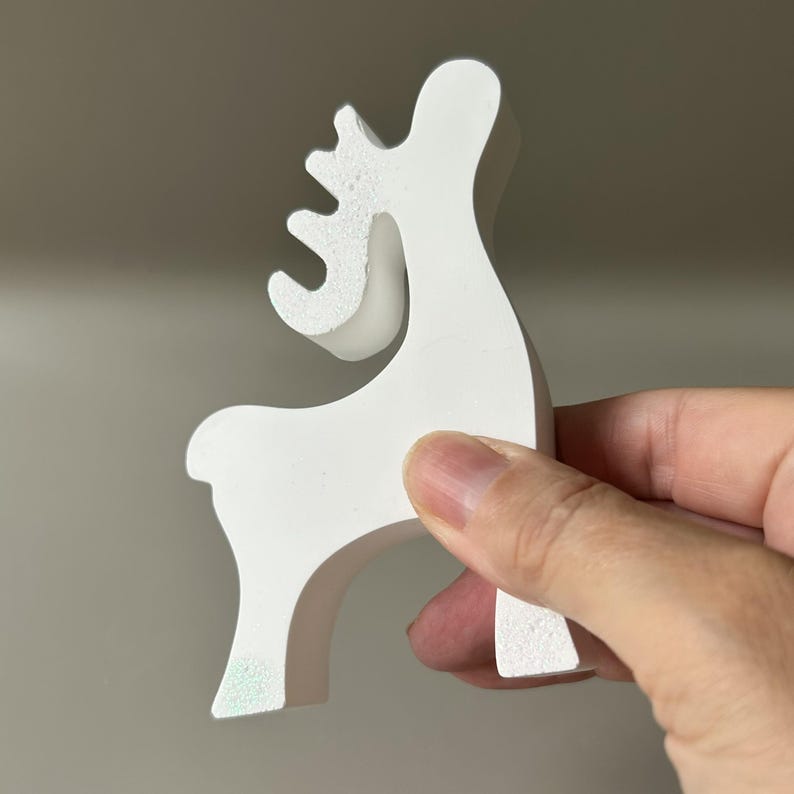 May include: A white, stylized reindeer figurine with a smooth, matte finish. The object features a simplified silhouette with a curved neck, a single antler branch, and a rounded tail, standing on two legs with a clean, minimalist aesthetic.