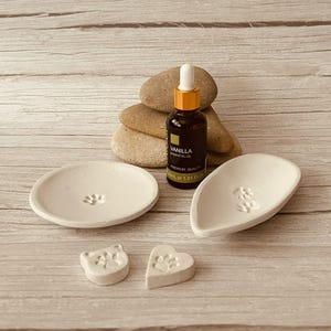 May include: A selection of white ceramic pet-themed items. Features two small dishes with paw prints, two paw print-shaped soaps, and a bottle of vanilla essential oil. The bottle is dark brown with gold detailing.