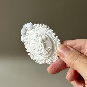 K&ouml;nnte beinhalten: Ein wei&szlig;es, ovales H&auml;ngeornament mit einem Relief einer gefl&uuml;gelten Feenfigur, umgeben von floralen Elementen. Das St&uuml;ck ist von einem verzierten Rand mit Schn&ouml;rkelmuster umrahmt und enth&auml;lt eine wei&szlig;e Bandschlaufe zum Aufh&auml;ngen.