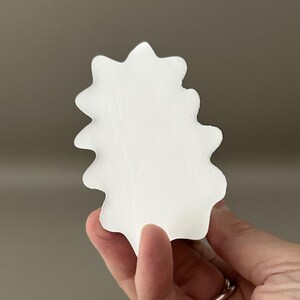 May include: A solid white, flat, leaf-shaped object with a smooth, matte surface and scalloped, wavy edges. The item features a symmetrical, organic silhouette with multiple rounded points along its perimeter, resembling a stylised botanical form.