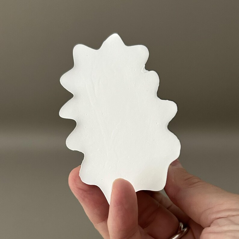 May include: A solid white, flat, leaf-shaped object with a smooth, matte surface and scalloped, wavy edges. The item features a symmetrical, organic silhouette with multiple rounded points along its perimeter, resembling a stylised botanical form.