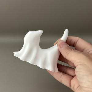 May include: A smooth, matte white figurine shaped like a stylised ghost with a curved, pointed tail and a draped, flowing body. The object features a rounded head and a hollowed-out section, presenting a solid, opaque appearance with clean lines.