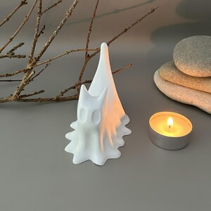 May include: A white, matte-finished figurine shaped like a stylised ghost with pointed ears and a draped, flowing base. The ghost figure has two hollow, oval-shaped eye sockets and a tall, tapered peak, standing upright as a decorative object.