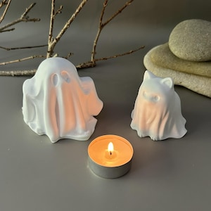 Może przedstawiać: Dwie białe, matowe figurki duch&oacute;w, jedna większa i jedna mniejsza z kocimi uszami, stoją obok małej, okrągłej świeczki typu tealight z płonącym pomarańczowym płomieniem. Figurki duch&oacute;w mają gładkie powierzchnie i proste wycięcia na oczy.