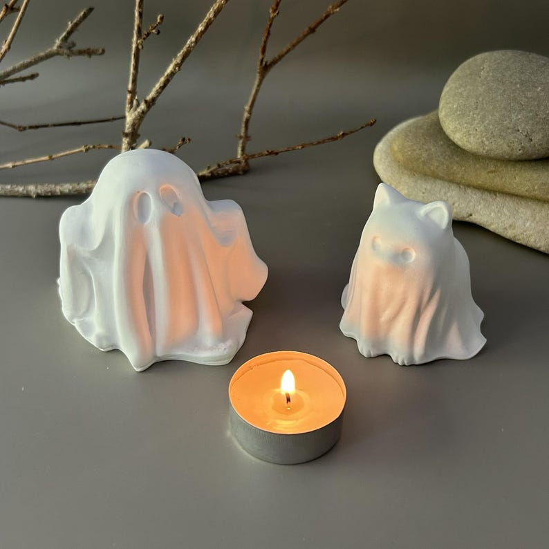 Może przedstawiać: Dwie białe, matowe figurki duch&oacute;w, jedna większa i jedna mniejsza z kocimi uszami, stoją obok małej, okrągłej świeczki typu tealight z płonącym pomarańczowym płomieniem. Figurki duch&oacute;w mają gładkie powierzchnie i proste wycięcia na oczy.