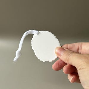 K&ouml;nnte beinhalten: Ein wei&szlig;es, oval geformtes Ornament mit gewelltem Rand und einer glatten, matten Oberfl&auml;che. Das Ornament verf&uuml;gt &uuml;ber eine wei&szlig;e elastische Kordel, die oben befestigt und am Ende verknotet ist, um es aufzuh&auml;ngen. Die Oberfl&auml;che ist schlicht, wei&szlig; und opak.
