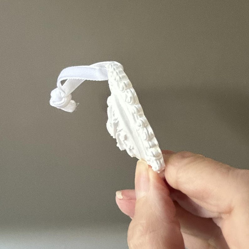 K&ouml;nnte beinhalten: Ein wei&szlig;es, kreisf&ouml;rmiges Ornament mit matter Oberfl&auml;che, das am &auml;u&szlig;eren Rand ein erhabenes, kompliziertes, schn&ouml;rkelartiges Reliefmuster aufweist. Das Ornament ist an einem d&uuml;nnen, wei&szlig;en Stoffband befestigt, das oben einfach verknotet ist.