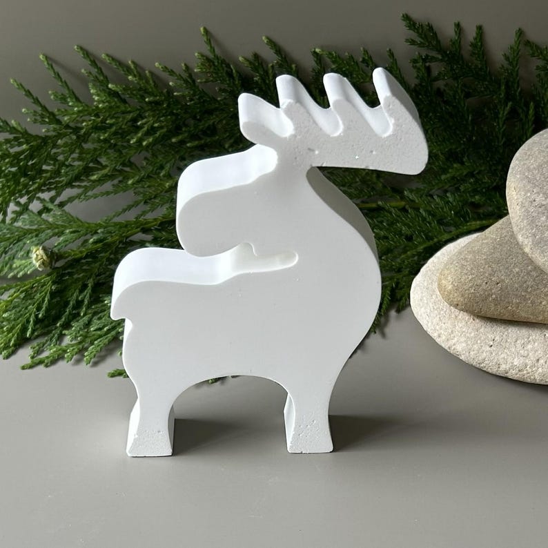 May include: A solid white, matte-finished figurine shaped like a stylized moose or reindeer in profile. The object features a smooth, continuous silhouette with a curved neck, a distinct antler structure, and four sturdy legs standing on a flat surface.