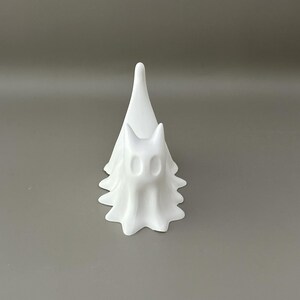 May include: A matte white figurine shaped like a stylised ghost with a pointed top and a scalloped base. The figure features two hollow, oval-shaped eye sockets and a smooth, continuous surface, standing upright with a draped, flowing appearance.