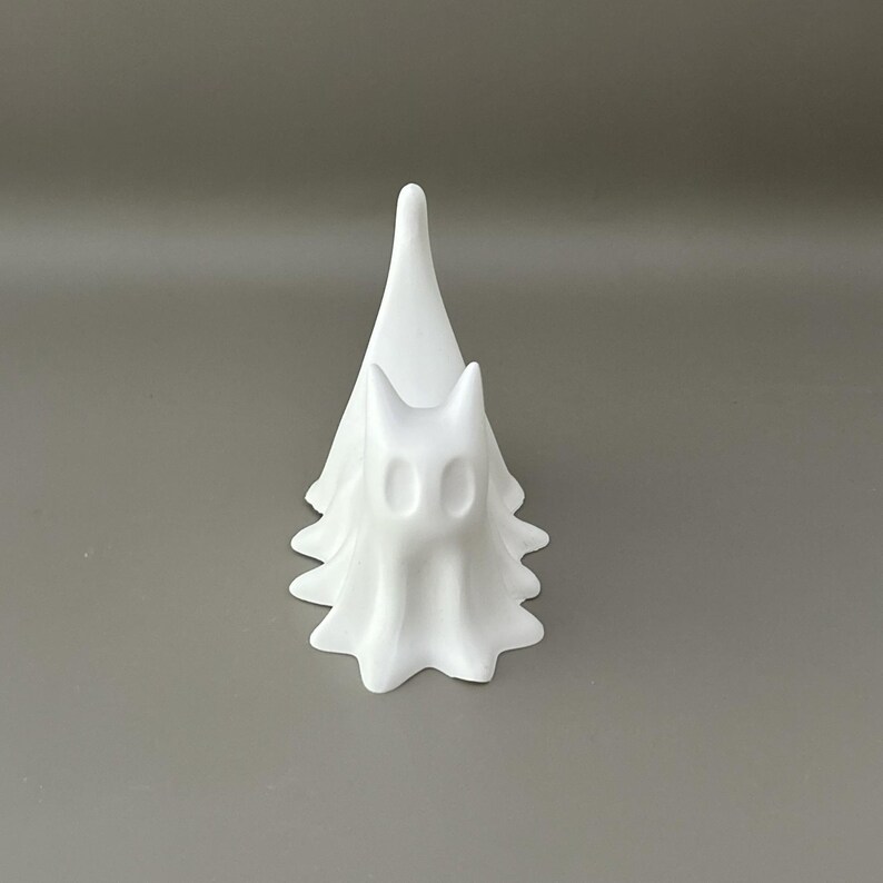 May include: A matte white figurine shaped like a stylised ghost with a pointed top and a scalloped base. The figure features two hollow, oval-shaped eye sockets and a smooth, continuous surface, standing upright with a draped, flowing appearance.