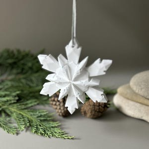 Pode incluir: Um ornamento de floco de neve branco com m&uacute;ltiplas facetas e uma estrutura geom&eacute;trica cristalina. O floco de neve apresenta pontas angulares afiadas e uma superf&iacute;cie texturizada, suspenso por uma fita fina de cor clara no topo central.