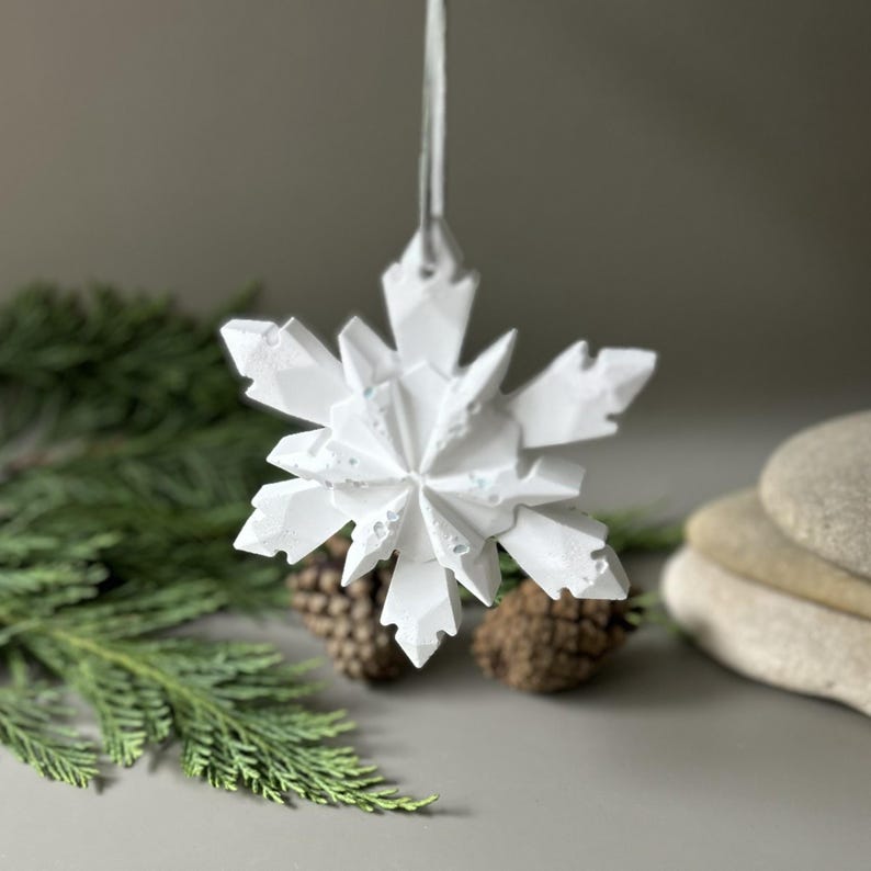 Pode incluir: Um ornamento de floco de neve branco com m&uacute;ltiplas facetas e uma estrutura geom&eacute;trica cristalina. O floco de neve apresenta pontas angulares afiadas e uma superf&iacute;cie texturizada, suspenso por uma fita fina de cor clara no topo central.
