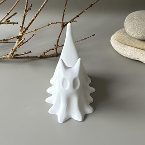 May include: A matte white ceramic ring holder shaped like a stylised ghost with a tall, pointed top and a wide, scalloped base. The figure features two hollow, oval-shaped eye cutouts on the front, providing a smooth, clean surface finish.