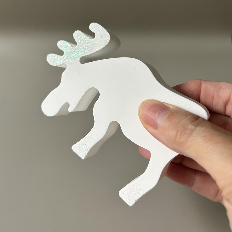 May include: A white, three-dimensional moose figurine crafted from a smooth material. The moose features a distinct, stylized antler design on its head that displays a subtle, shimmering iridescent or holographic glitter finish across the surface.