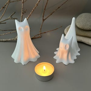 May include: Two white ghost-shaped figurines with smooth, flowing forms and hollow eye sockets, positioned next to a small, round metal tea light candle holder containing a lit candle with a warm, glowing yellow flame.